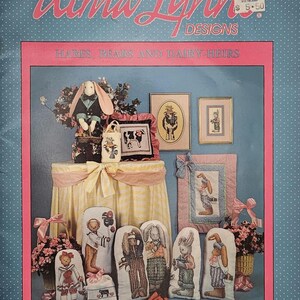 May include: A catalog cover featuring cross-stitch designs. The title "Alma Lynn Designs" is in pink script. The image showcases framed cross-stitch pieces, including rabbits and bears, and decorative pillows. The text "HARES, BEARS AND DAIRY-HEIRS" is displayed.