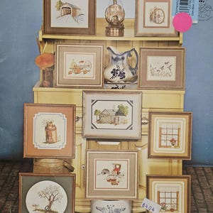 May include: A collection of framed cross-stitch samplers with rustic themes, including barns, a water wheel, and a milk can. The display also features a decorative oil lamp and a blue and white pitcher. The text "RUSTIC REFLECTIONS" is visible.