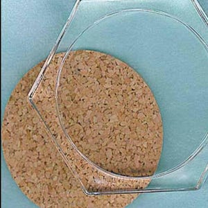May include: A clear, hexagonal glass coaster sits atop a round cork coaster. The cork coaster is placed on a light blue surface. The glass coaster has a simple, modern design.