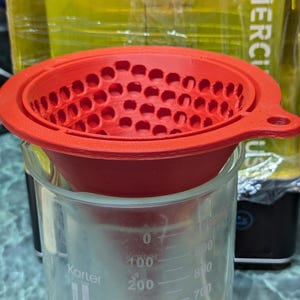 Può includere: Un imbuto in silicone rosso con interno perforato è posto sopra un becher di vetro trasparente. Il becher ha segni di graduazione e il nome "Karter" stampato sul lato. L'imbuto è progettato per filtrare o versare liquidi.