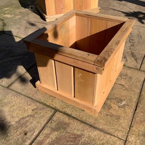 May include: Two unfinished wooden planters with a square shape. The planters are made of light-colored wood and have a simple, modern design. The planters are sitting on a stone surface.