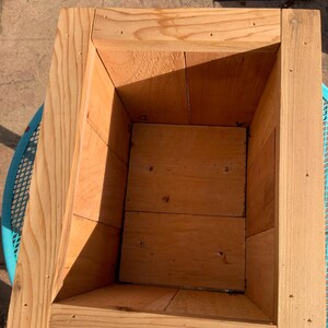K&ouml;nnte beinhalten: Ein rechteckiger Pflanzkasten aus Holz mit nat&uuml;rlicher Oberfl&auml;che. Der Kasten hat einen sich verj&uuml;ngenden Innenraum und besteht aus mehreren Holzst&uuml;cken. Der Pflanzkasten steht auf einem blauen Metalltisch.