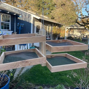 Könnte beinhalten: Ein hängender Vogelhaus-Futterspender aus hellem Holz mit vier quadratischen Tabletts mit grünen Maschenböden. Der Futterspender wird an einer Kette aufgehängt. Das Holz hat ein natürliches, unbehandeltes Aussehen. Jedes Tablett ist etwa 30 cm groß.