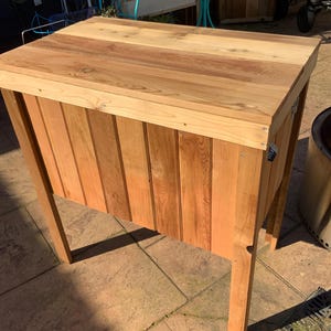 Könnte beinhalten: Eine rechteckige Kühlbox aus Holz mit vertikalen Zedernholzplanken, einem flachen Deckel und vier stabilen Beinen. Die Außenseite zeigt eine natürliche hellbraune Holzmaserung mit sichtbaren Metallschrauben am oberen Rahmen und einem Flaschenöffner.