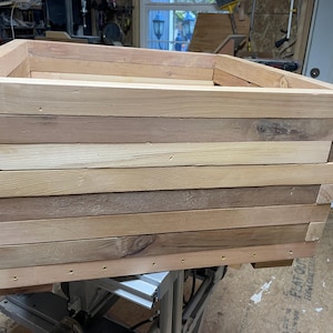 May include: A rectangular wooden planter box constructed from layered, light-colored wood planks. The box has a simple, open design, with visible wood grain and natural variations in color. The planter is ready for plants.