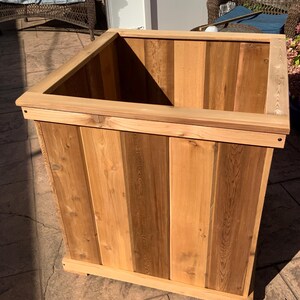 May include: A square wooden planter box constructed from vertical cedar planks with a flat top rim. The natural wood features varying shades of light and medium brown grain patterns, with a sturdy base and a hollow interior for holding plants.