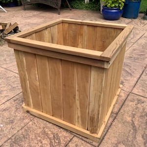 May include: A large, square wooden planter box made of light brown wood. The planter has a rectangular shape with a decorative top trim and base. The wood grain is visible, and the box appears to be empty.