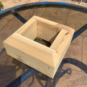 Könnte beinhalten: Eine quadratische Pflanzkasten aus hellem Holz. Die Box besteht aus mehreren Holzstücken, die mit Messingschrauben an den Seiten verbunden sind. Der Pflanzkasten hat einen hohlen Innenraum und eine abgeschrägte Oberkante.