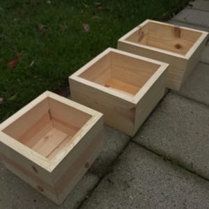May include: Three unfinished wooden square planters. The planters are made of light-colored wood and have a simple, boxy design. They are arranged in a row on a gray stone surface, with green grass visible in the background. The planters are empty.