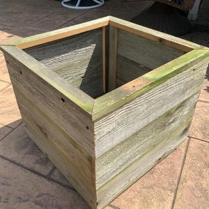 May include: A square planter box constructed from weathered gray wooden planks with visible wood grain and green-tinted edges. The container features a mitered top rim and a hollow interior, showcasing a rustic, unfinished appearance with exposed nail heads.