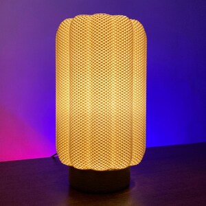 May include: A cylindrical table lamp with a textured, woven-like shade. The lamp emits a warm, soft glow. The base is a dark, solid color. The background is a gradient of purple and blue.