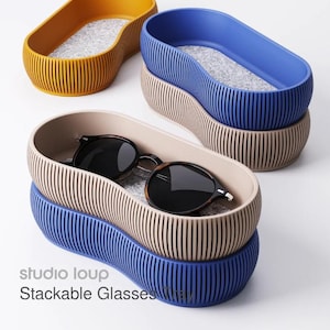 May include: Stackable glasses trays in mustard yellow, blue, and beige. Each tray has a ribbed exterior and a felt interior. One tray holds a pair of sunglasses. The text "studio loup Stackable Glasses" is visible.