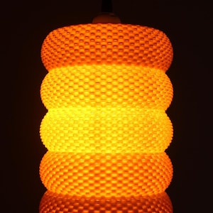 May include: A hanging, cylindrical lamp with a textured, honeycomb-like surface. The lamp emits a warm, orange glow, creating a cozy ambiance. The design features three stacked, rounded sections, each with a detailed pattern. The background is dark.