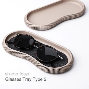 May include: A beige oval glasses tray with a ribbed edge, holding a pair of sunglasses with dark lenses. The tray has a dark gray felt lining. The text "studio loup Glasses Tray Type 3" is visible.