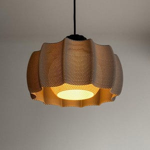 May include: A beige, textured pendant light fixture with a unique, wavy design. The light hangs from a black cord and fixture. The interior of the lamp is illuminated with a warm, inviting glow, creating a soft ambiance. The lamp's dimensions are not visible.