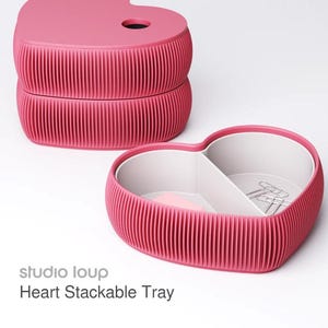 May include: Pink heart-shaped stackable tray with a ribbed exterior and a white interior. The top tray has a hole. The text "studio loup Heart Stackable Tray" is visible.