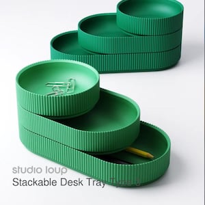 May include: Green stackable desk trays with a ribbed texture. The trays are in a stepped arrangement, with paperclips and a pencil visible in the trays. The text "studio loup Stackable Desk Tray Type 0" is printed on the bottom left.
