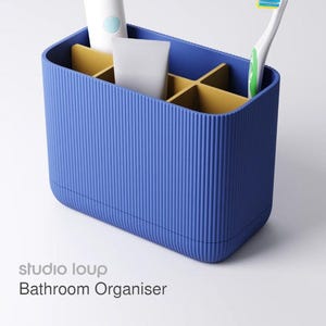 May include: A blue bathroom organizer with vertical ribbed texture. The organizer has multiple compartments with a yellow interior. It holds a toothbrush, toothpaste, and a tube of product. The text "studio loup Bathroom Organiser" is visible.