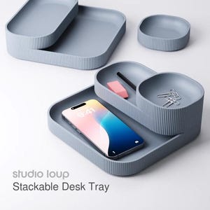 May include: A set of stackable desk trays in a light blue-grey color. The trays have a ribbed texture and various shapes, including rectangular and circular. A smartphone, pencil, eraser, and paper clips are inside the trays. The text "studio loup Stackable Desk Tray" is visible.