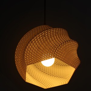 May include: A woven, geometric pendant light fixture with a warm, golden hue. The light has a unique, textured surface and a bright, spherical bulb at its center. The fixture is suspended from a black cord against a dark background.