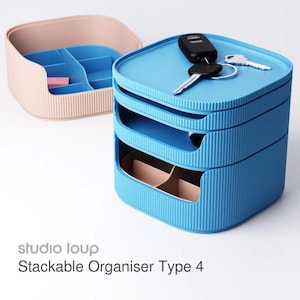 May include: A blue and tan stackable organizer with a ribbed texture. The organizer has multiple compartments and drawers. Keys and a key fob rest on top. The text "studio loup Stackable Organiser Type 4" is visible.
