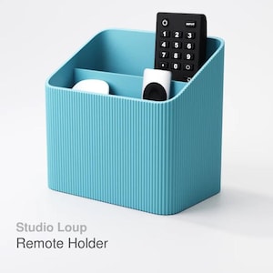 May include: A teal blue remote holder with vertical ribbed texture. The holder contains a black remote control, a white device, and a small white object. The text "Studio Loup Remote Holder" is visible.