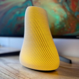 May include: A yellow, textured lamp with a unique, curved shape. The lamp has a woven pattern and emits a warm, soft glow. The lamp is sitting on a wooden surface.
