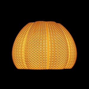 May include: A woven, dome-shaped lampshade in a warm, golden-yellow hue. The shade has a textured surface with a repeating pattern of small, rectangular openings. The background is solid black, highlighting the shade's shape and color.