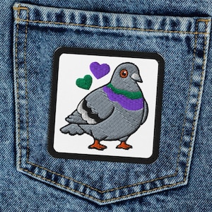May include: A square patch featuring a cartoon pigeon with a purple and green design, set against a white background. The patch is sewn onto the back pocket of a pair of blue denim jeans.