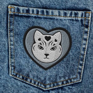 May include: Light blue denim jeans with a cat-shaped patch on the back pocket. The patch features a gray cat face inside a gray heart with a black border. The jeans have a classic five-pocket design.