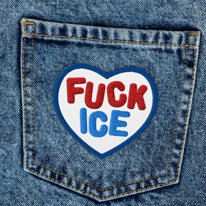 Könnte beinhalten: Nahaufnahme einer blauen Jeans mit einem herzförmigen Aufnäher auf der Gesäßtasche. Der Aufnäher hat einen weißen Hintergrund mit einem rot-blauen Design mit der Aufschrift "FUCK ICE". Die Jeans hat ein klassisches Fünf-Taschen-Design.