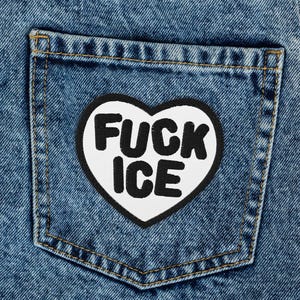Könnte beinhalten: Nahaufnahme einer blauen Jeans mit einem weißen herzförmigen Aufnäher auf der Gesäßtasche. Der Aufnäher hat schwarzen Text mit der Aufschrift "FUCK ICE". Die Jeans ist gefaltet und zeigt die Textur und Waschung des Denims.