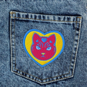 May include: Close-up of acid wash denim jeans with a cat-shaped patch on the back pocket. The patch features a pink cat face inside a yellow heart with a blue border. The jeans have a classic five-pocket design.