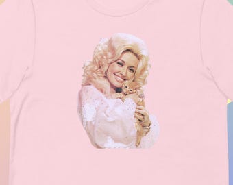 May include: A light pink short-sleeved t-shirt featuring a centered graphic print of a person with blonde hair holding a small orange kitten. The person is wearing a white long-sleeved garment with lace details and is smiling at the camera.