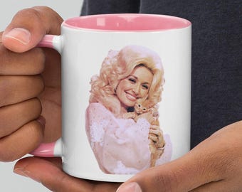 Puede incluir: Taza de cerámica blanca con interior y asa rosas. La taza presenta un retrato de una mujer sonriente con cabello rubio sosteniendo un pequeño animal. La taza está sostenida por una persona.