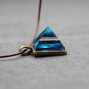 May include: A pyramid-shaped pendant with a blue and black layered design. The pendant is set in a bronze-colored frame and hangs from a brown cord. The pyramid has a geometric shape and is a unique piece of jewelry.