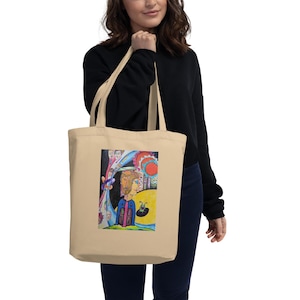 May include: Cream-coloured tote bag displaying a vibrant, abstract artwork. The illustration presents a surreal scene with figures, a sun, and diverse shapes. The bag has long handles and is held by a person wearing a black top and blue jeans.