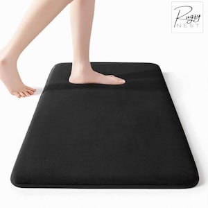 May include: A black rectangular bath mat with rounded corners. A person's foot is stepping onto the mat. The Rugsy Nest logo is in the upper right corner.