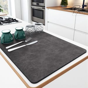 May include: A dark grey rectangular placemat with a textured surface. Two green mugs, two clear glasses, a fork, and a knife are arranged on the mat. The scene is set on a white countertop in a kitchen.