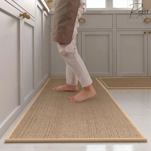 Peut inclure: Un tapis de cuisine rectangulaire beige tissé avec une bordure beige clair. Le tapis est posé sur un sol carrelé blanc, avec un plus petit tapis assorti visible en arrière-plan. L'image est prise dans une cuisine.