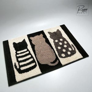 Può includere: Un zerbino rettangolare con tre sagome di gatti in nero, marrone e grigio scuro. I gatti sono posti su uno sfondo a strisce nere e bianco sporco. Lo zerbino ha un bordo nero. Il marchio "Rugsy Nest" è nell'angolo in alto a destra.