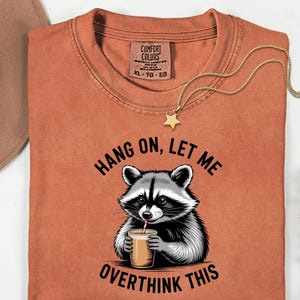 May include: A rust-coloured t-shirt featuring a raccoon graphic drinking from a cup, with the text "HANG ON, LET ME OVERTHINK THIS." A gold necklace with a star pendant is draped on the shirt. A brown hat is visible in the background.