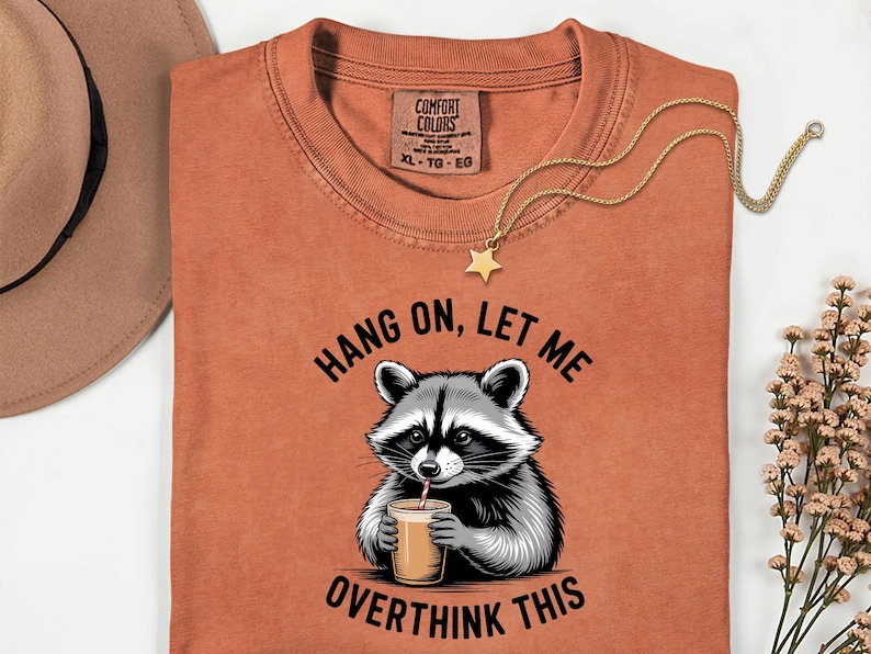 May include: A rust-coloured t-shirt featuring a raccoon graphic drinking from a cup, with the text "HANG ON, LET ME OVERTHINK THIS." A gold necklace with a star pendant is draped on the shirt. A brown hat is visible in the background.