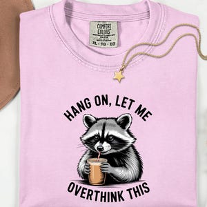 May include: Pale pink t-shirt featuring a raccoon illustration and the phrase "HANG ON, LET ME OVERTHINK THIS". A gold-coloured star pendant necklace is draped across the shirt.