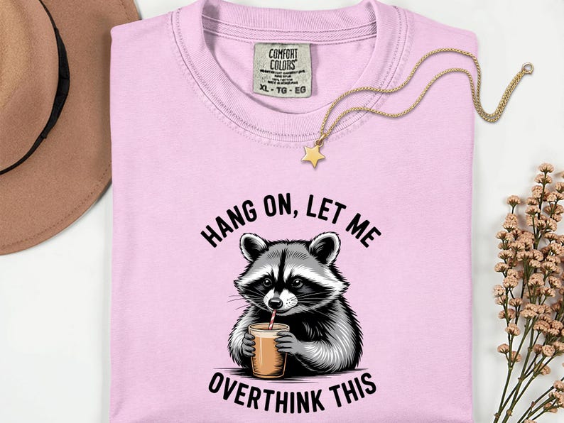 May include: Pale pink t-shirt featuring a raccoon illustration and the phrase "HANG ON, LET ME OVERTHINK THIS". A gold-coloured star pendant necklace is draped across the shirt.