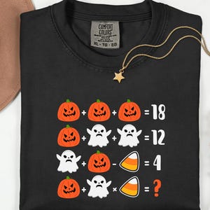 May include: Black Comfort Colors XL-TG-EG t-shirt featuring a Halloween-themed math problem. The graphic includes orange pumpkins, white ghosts, and candy corn, with equations and a question mark.