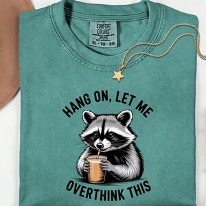 May include: Teal Comfort Colors t-shirt featuring a raccoon graphic drinking from a cup. The text reads "Hang on, let me overthink this." A gold star pendant necklace and a brown hat are also visible.