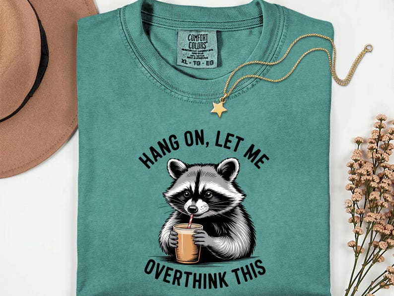 May include: Teal Comfort Colors t-shirt featuring a raccoon graphic drinking from a cup. The text reads "Hang on, let me overthink this." A gold star pendant necklace and a brown hat are also visible.