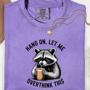 May include: A lilac Comfort Colors t-shirt with a raccoon graphic drinking from a cup, and the text "Hang on, let me overthink this." A gold necklace with a star pendant is draped on the shirt.