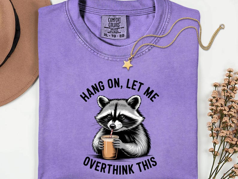 May include: A lilac Comfort Colors t-shirt with a raccoon graphic drinking from a cup, and the text "Hang on, let me overthink this." A gold necklace with a star pendant is draped on the shirt.